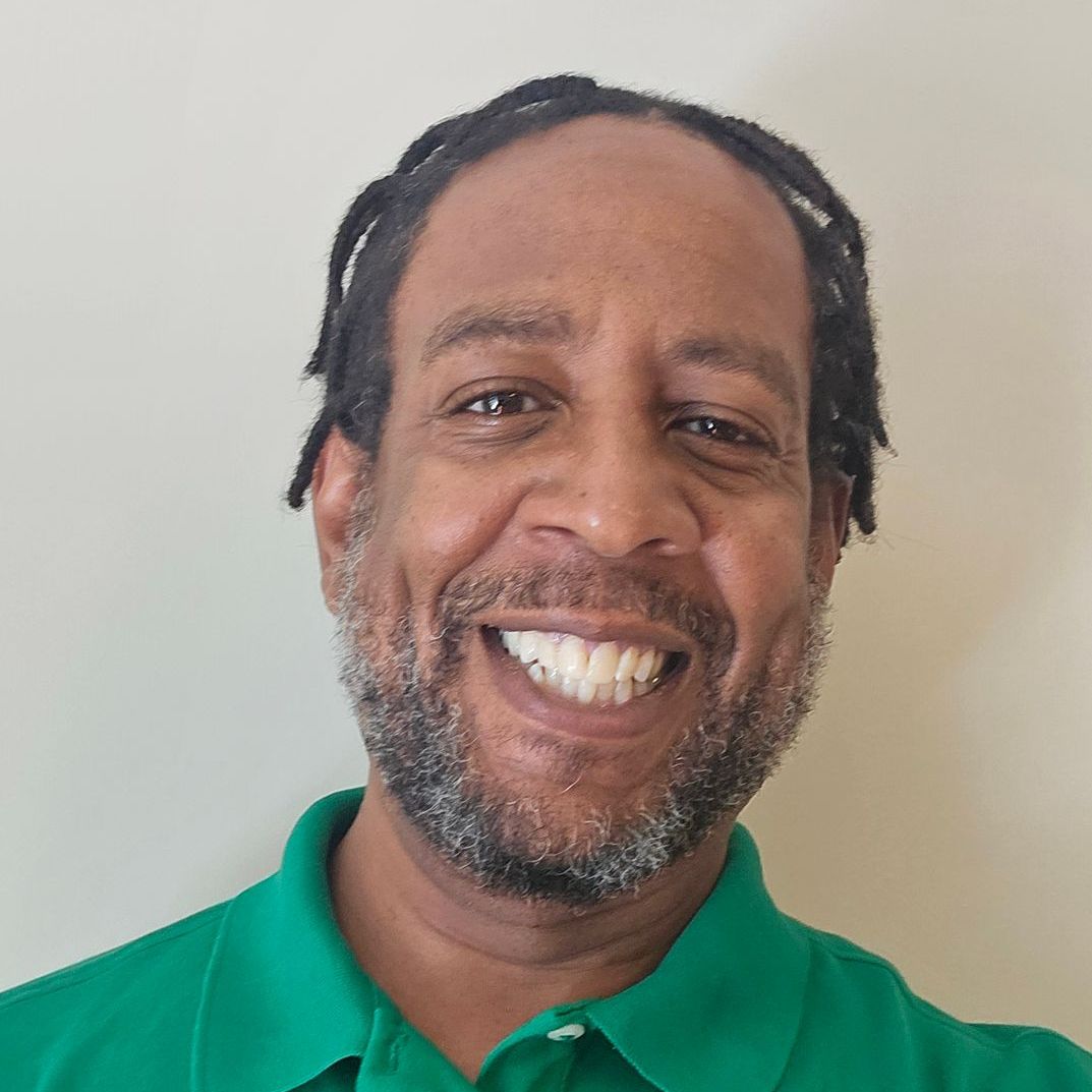 A man with dreadlocks and a beard is wearing a green polo shirt and smiling.