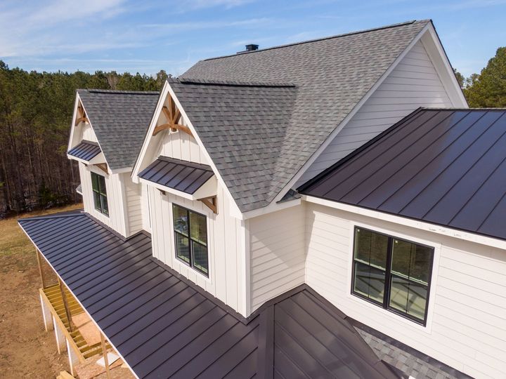 White farmhouse with gray shingle roof, black metal roof accents, and forest backdrop.