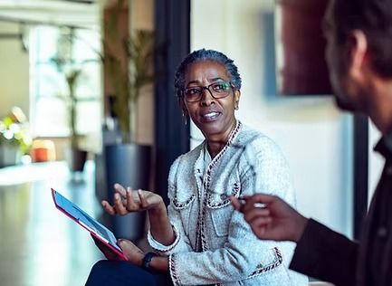 Woman in blazer gestures while speaking to a man; she holds a tablet.