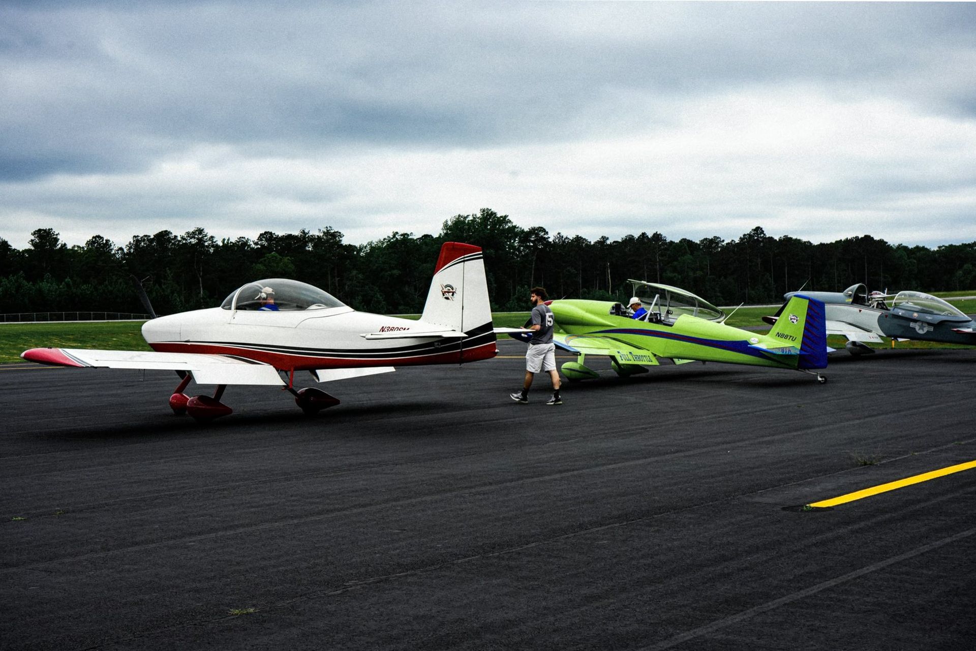 Planes on an asphalt runway; pilot stands next to a green plane; red and white plane is closest. Cloudy sky.
