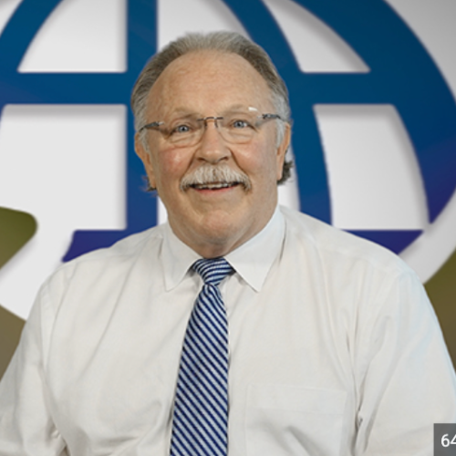 Smiling older man in glasses, white shirt, blue striped tie, in front of a blue globe logo.