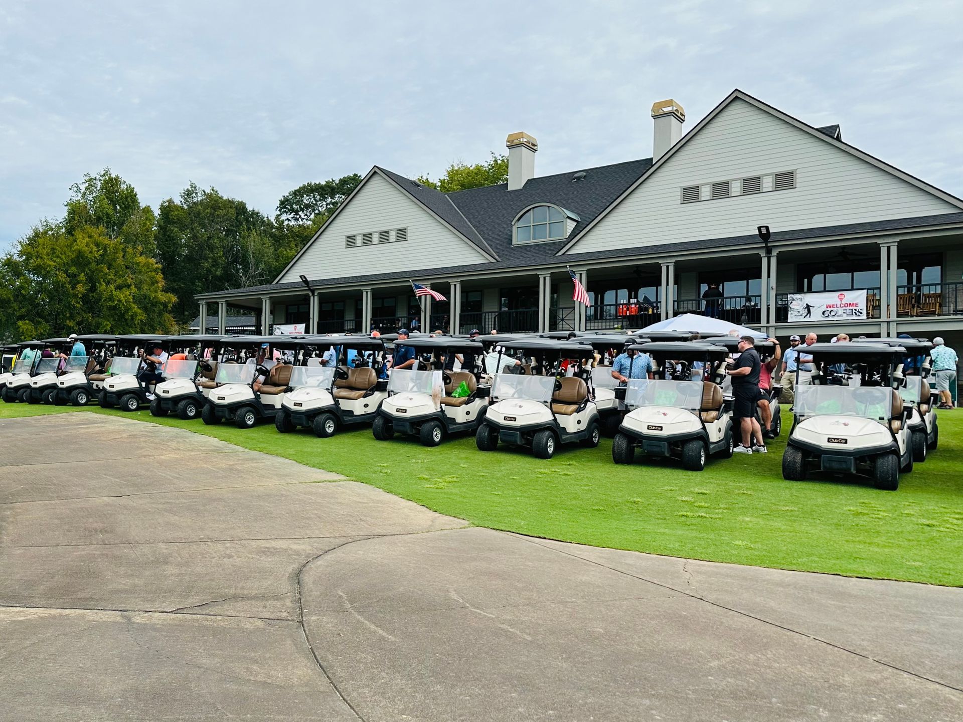 Golf carts lined up on grass in front of a clubhouse; people are standing around.
