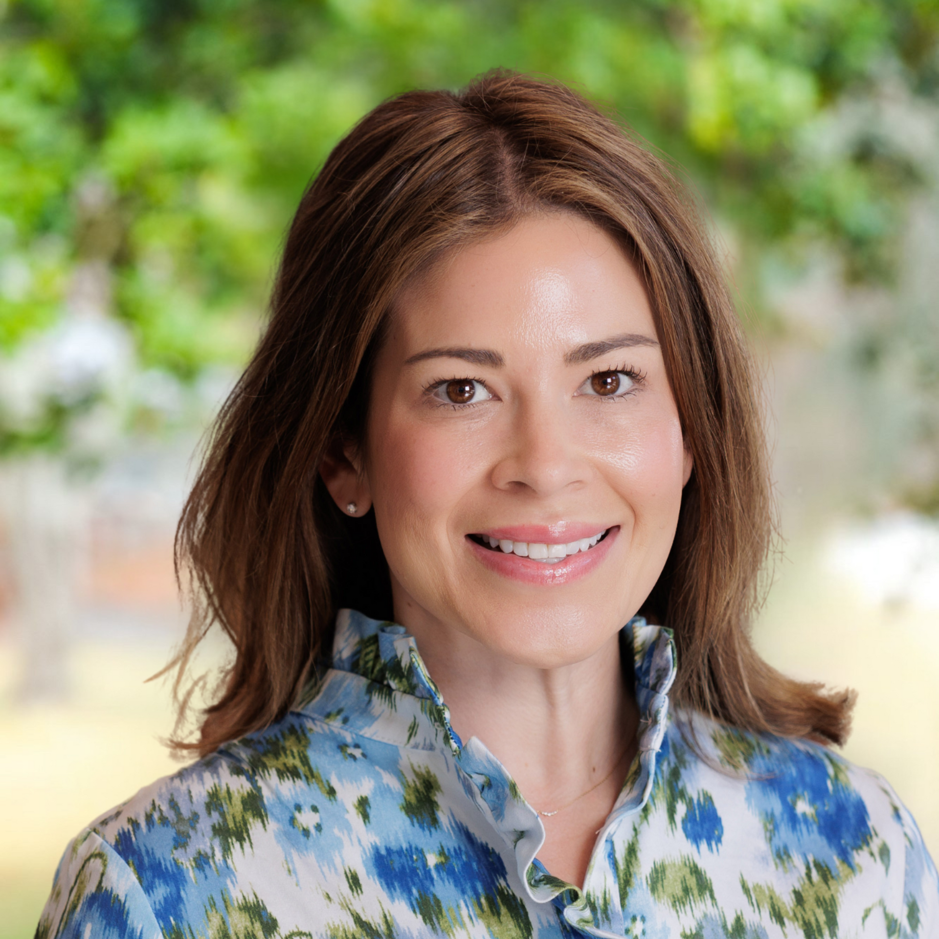 Woman with brown hair smiling, wearing blue floral shirt, against a blurred green background.