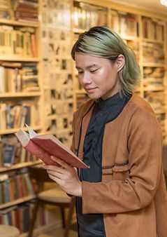 Person with green-tinted hair reads a book in a library, wearing a brown jacket and black shirt.