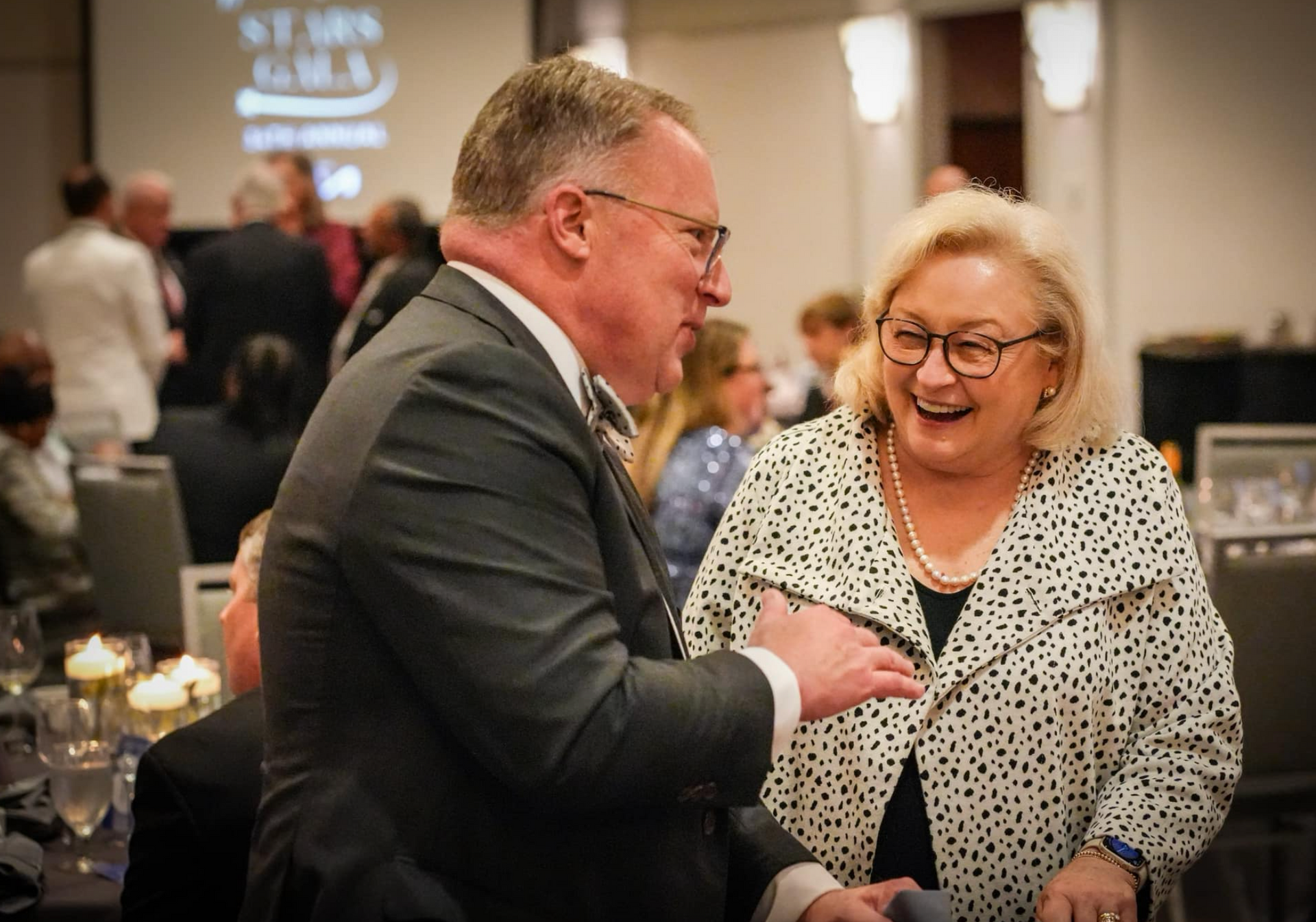 Man in suit chats with smiling woman in polka dot jacket at a formal event.