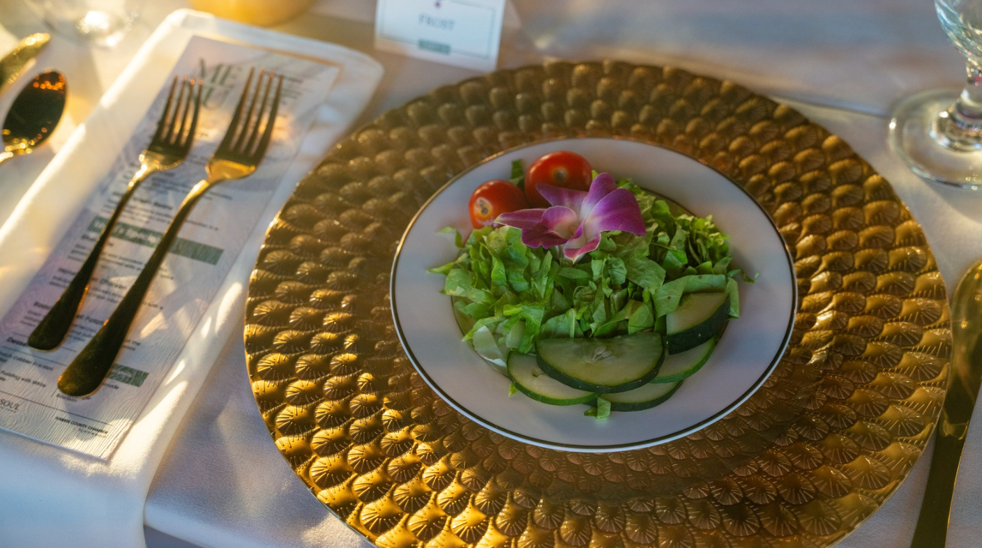 Salad on a white plate, gold charger, with fork and knife on a white table, outdoors.