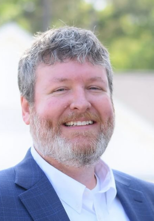 Smiling man with graying beard, in blue suit, white shirt. Outdoor setting.