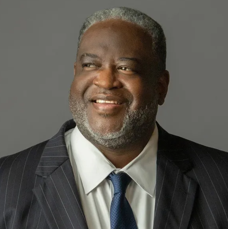 Mature Black man in suit, smiling and looking right, gray background.