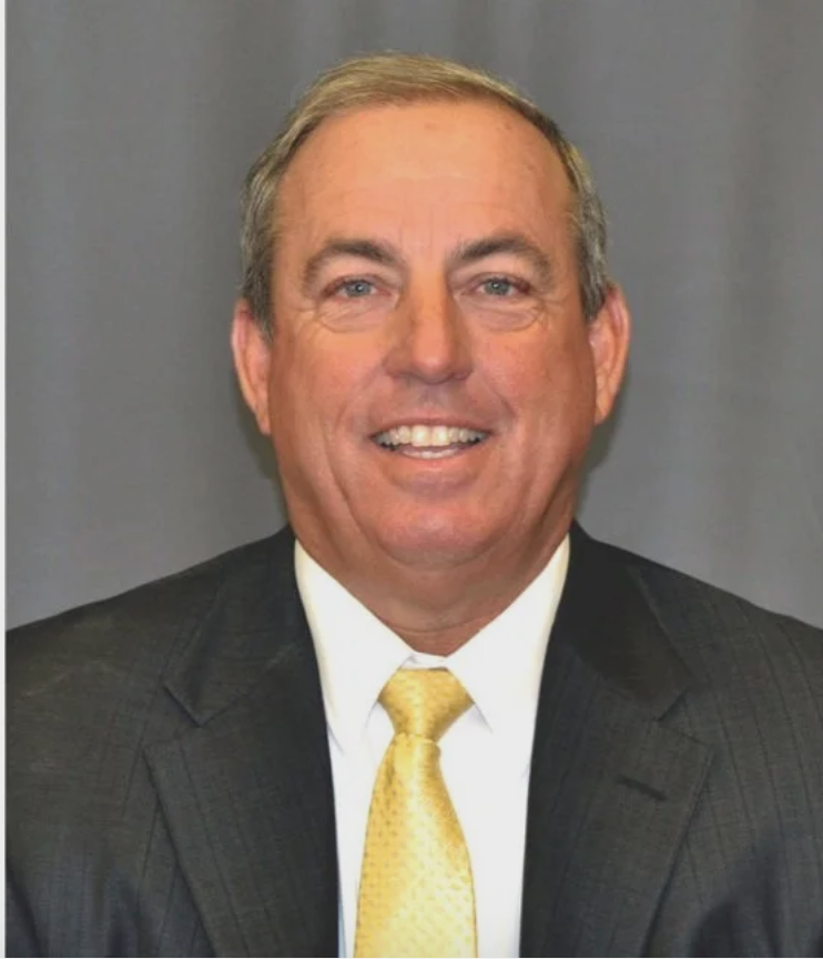 Man in a suit smiles at the camera. He wears a yellow tie. Gray background.