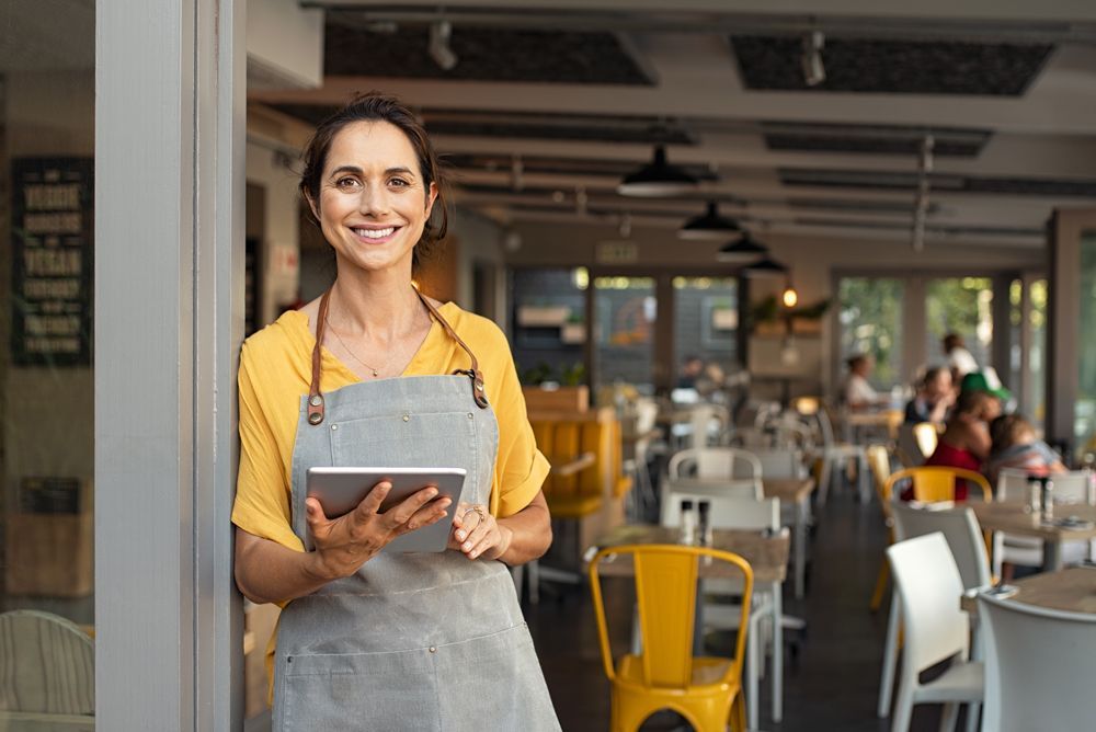 a woman in an apron holds a tablet in front of a restaurant