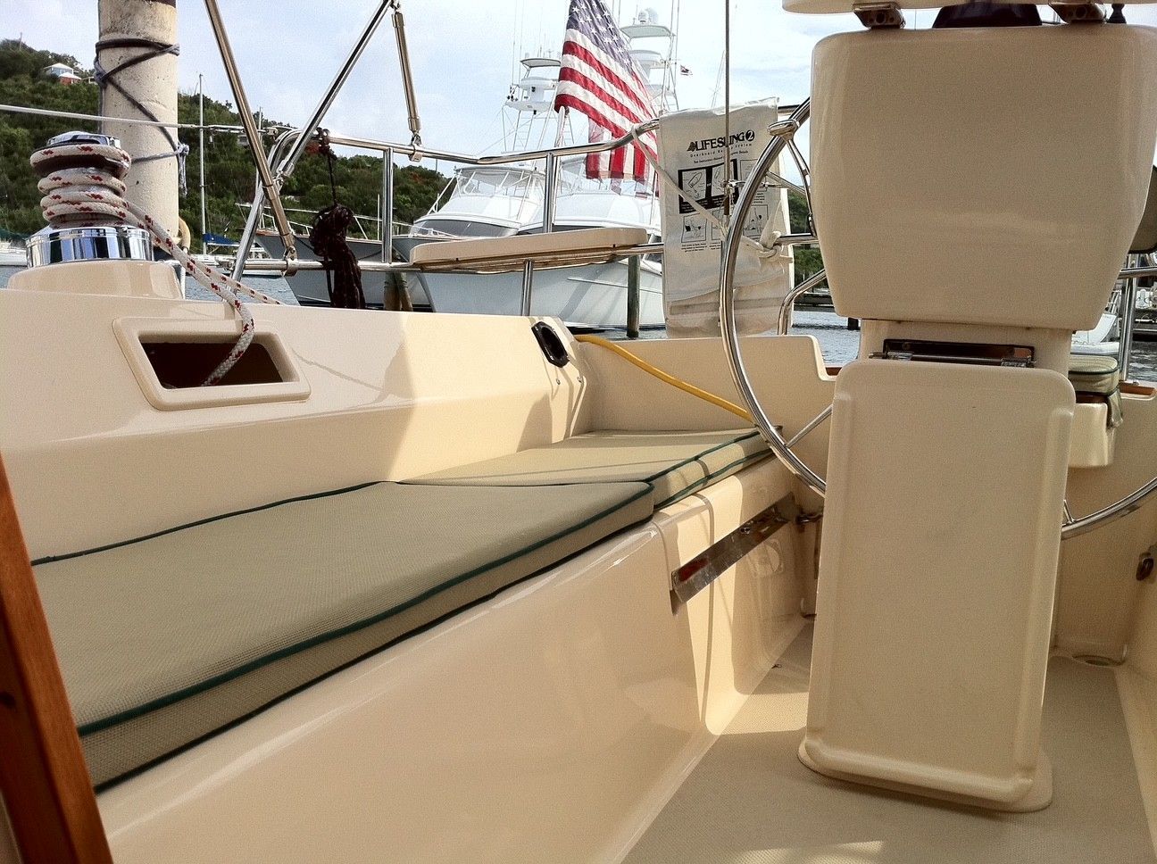 View of a sailboat deck with a steering wheel and an American flag in the background. Beige deck, green cushions.