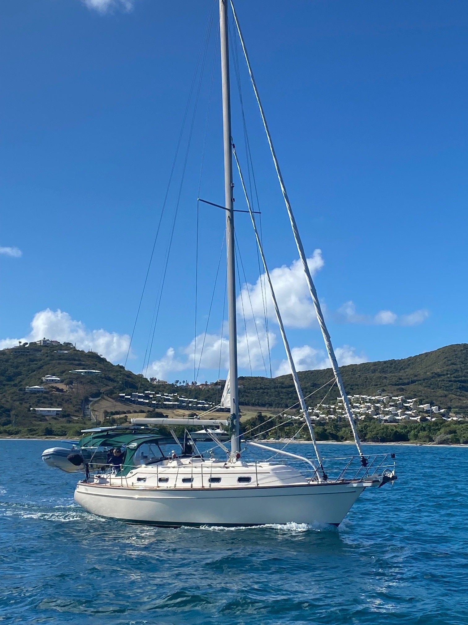 Sailboat on blue water with a green-covered cockpit, under a blue sky, near a hilly shoreline.