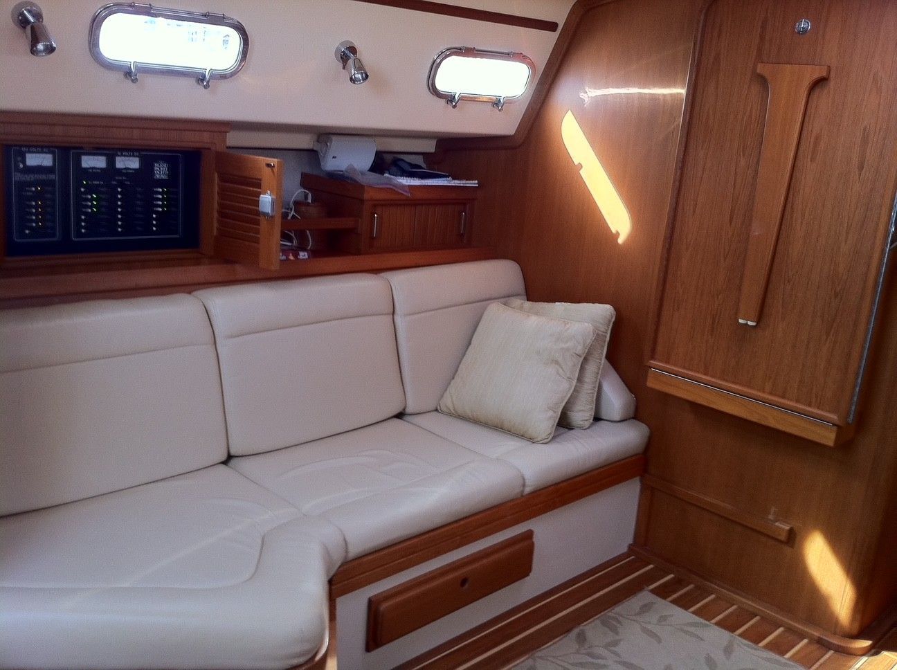 Interior of a sailboat: beige couch, wooden paneling, small windows, and a control panel.