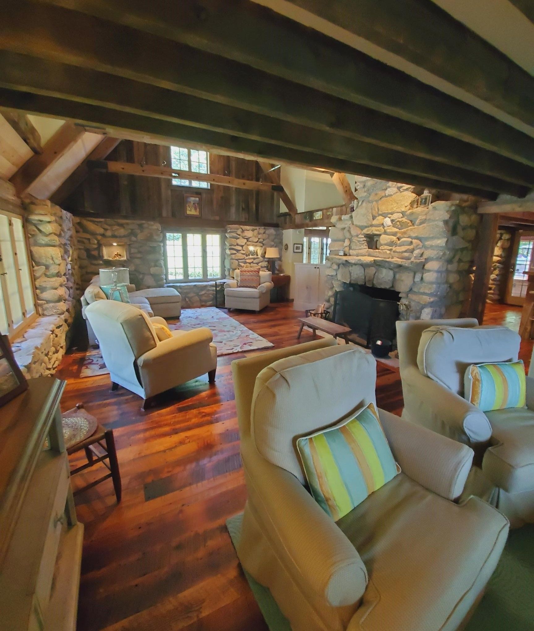 Cozy living room with stone fireplace, wooden beams, hardwood floors, and comfortable cream-colored armchairs.