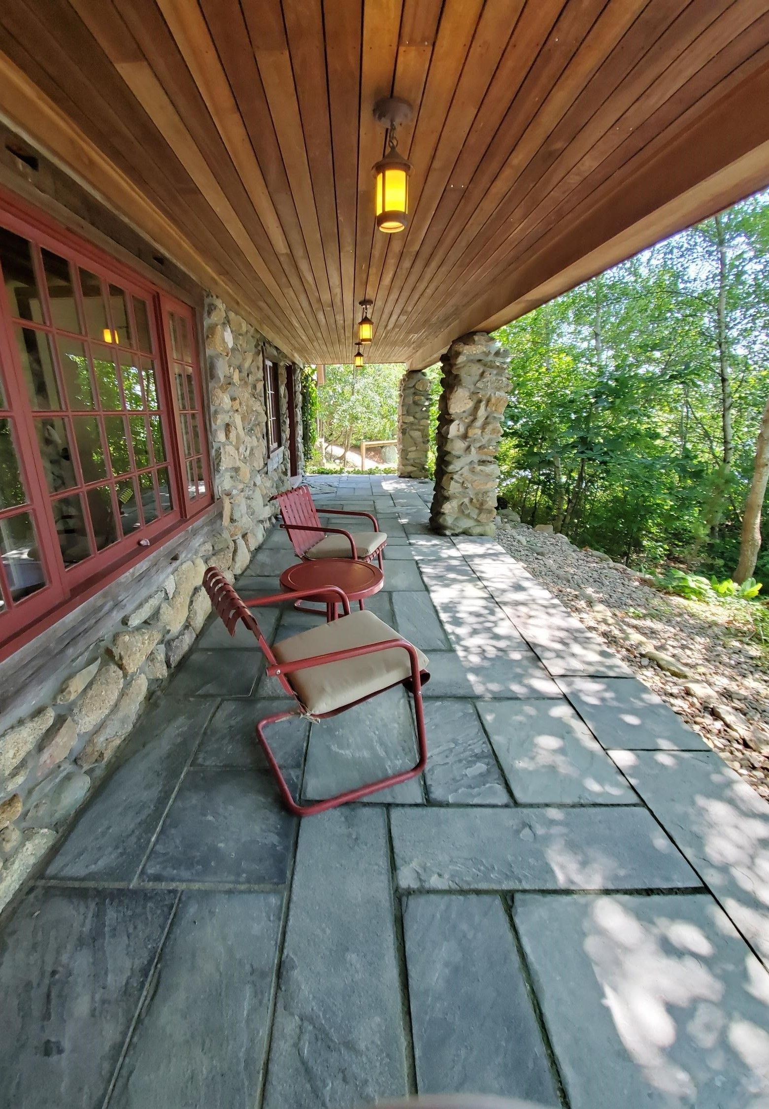 Stone porch with red-framed chairs, stone floor, and wood ceiling; view of trees in background.