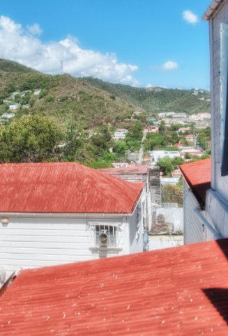 A view from above looking down on buildings with red roofs towards a green, hilly landscape under a blue sky.