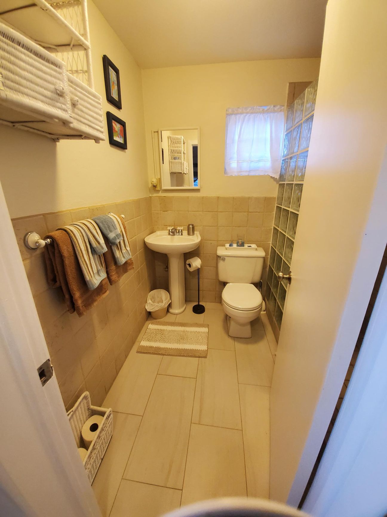 Small bathroom with toilet, sink, glass block wall, towels, and shelves.