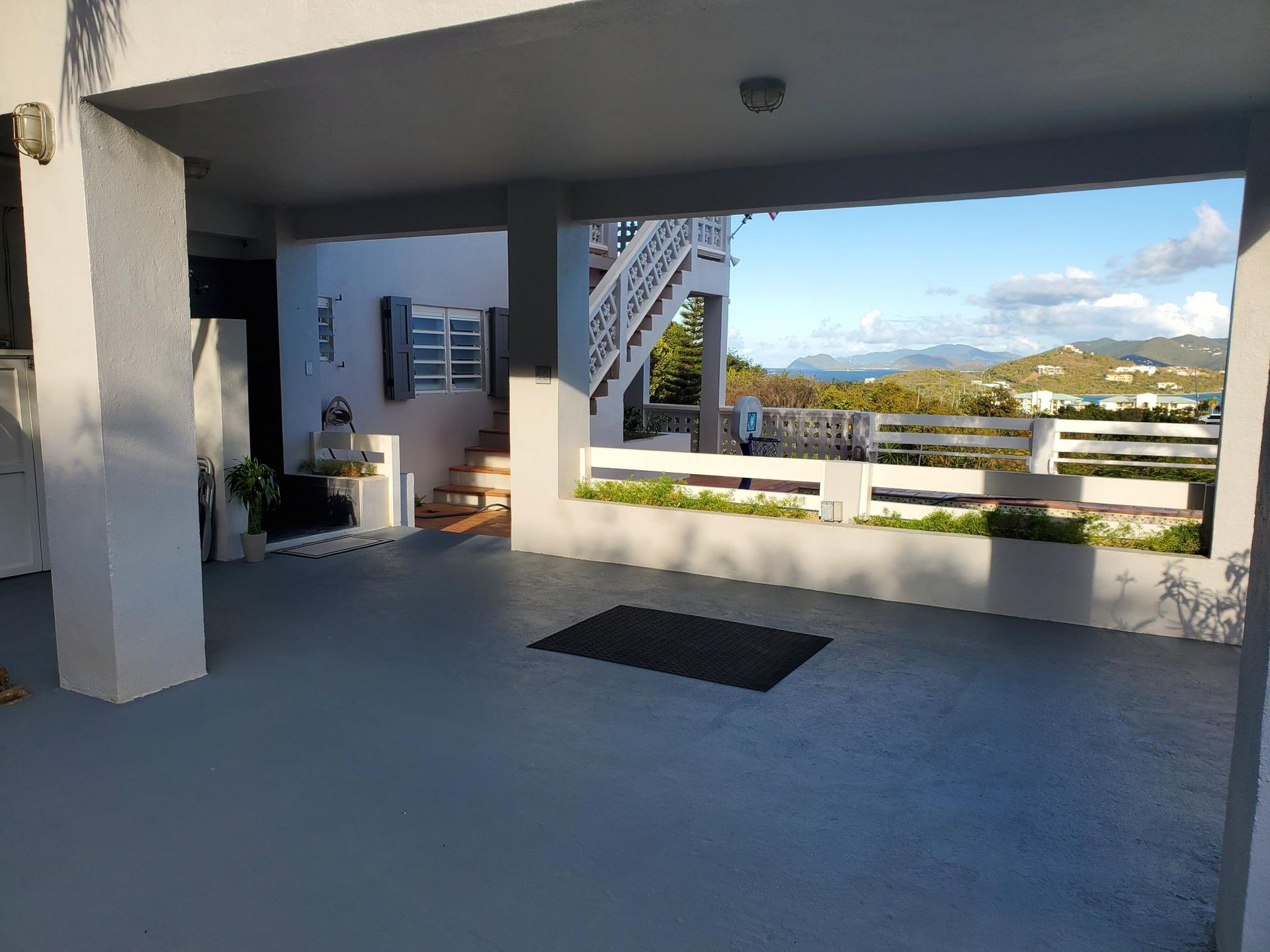 Covered outdoor patio with a view of the sea and islands. White pillars, gray floor, and blue sky.