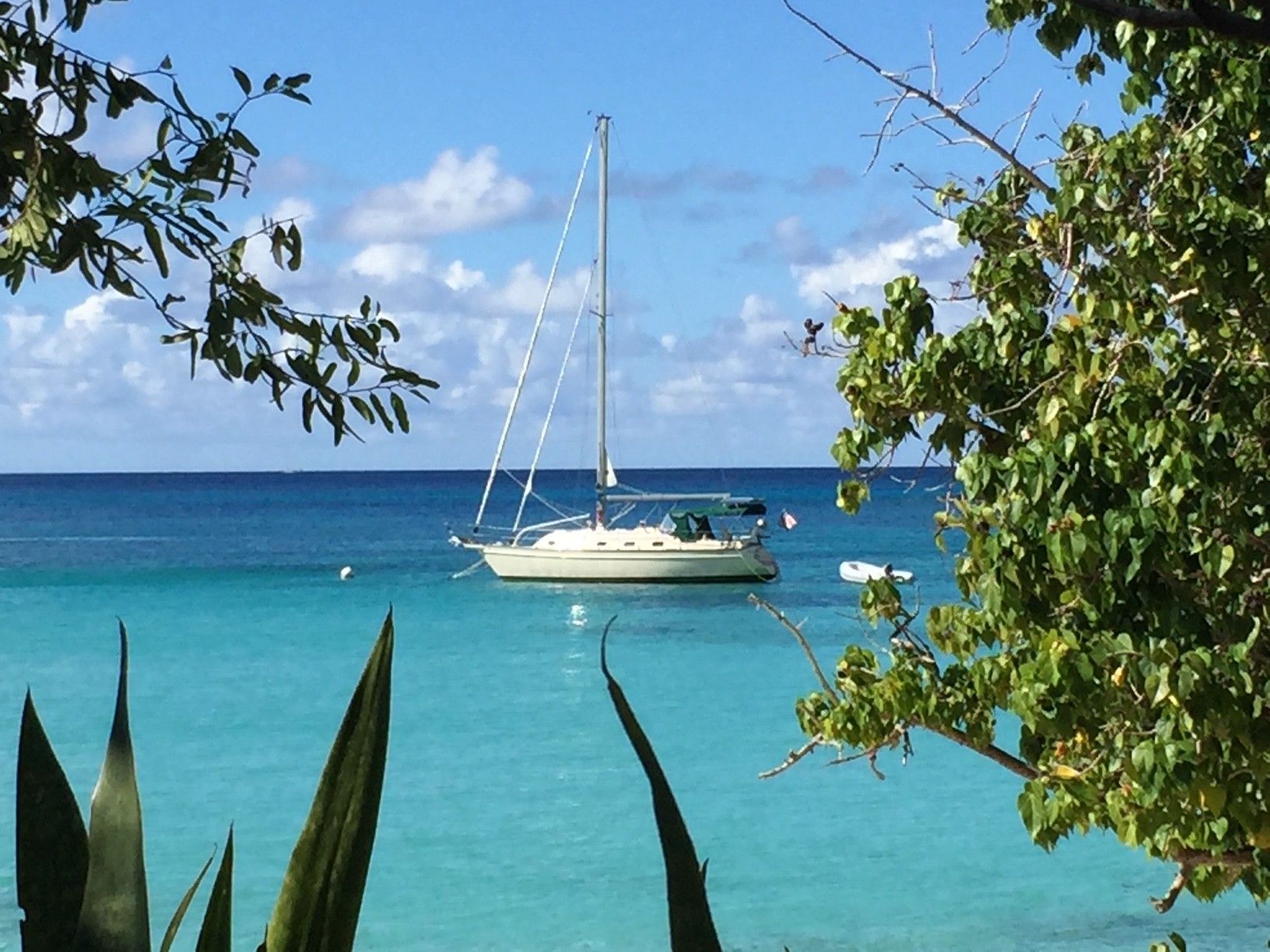 Sailboat anchored in turquoise water, framed by green foliage, under a blue sky with clouds.