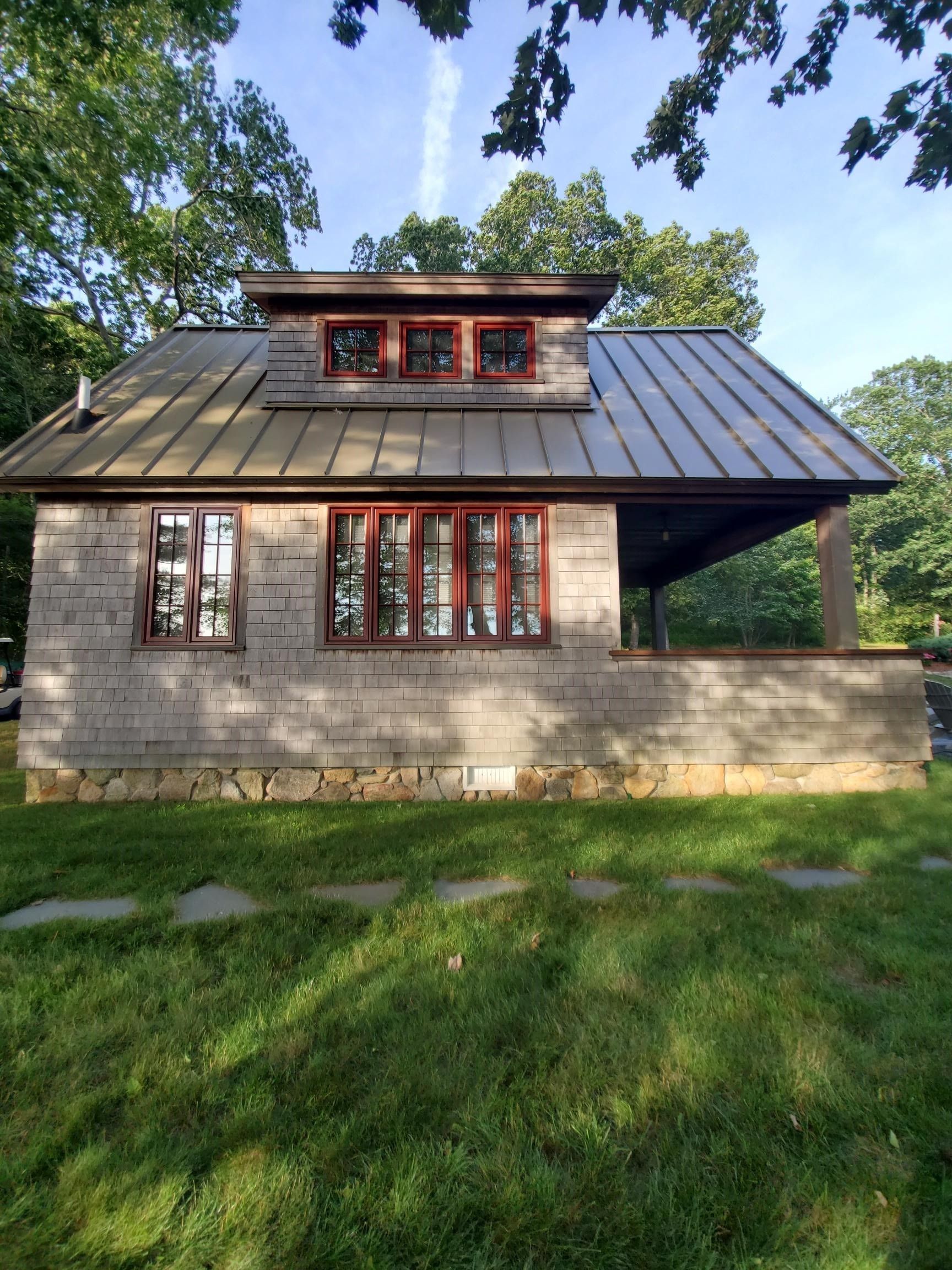 Small wood-shingled cottage with red-framed windows and metal roof, under trees, with stone path.