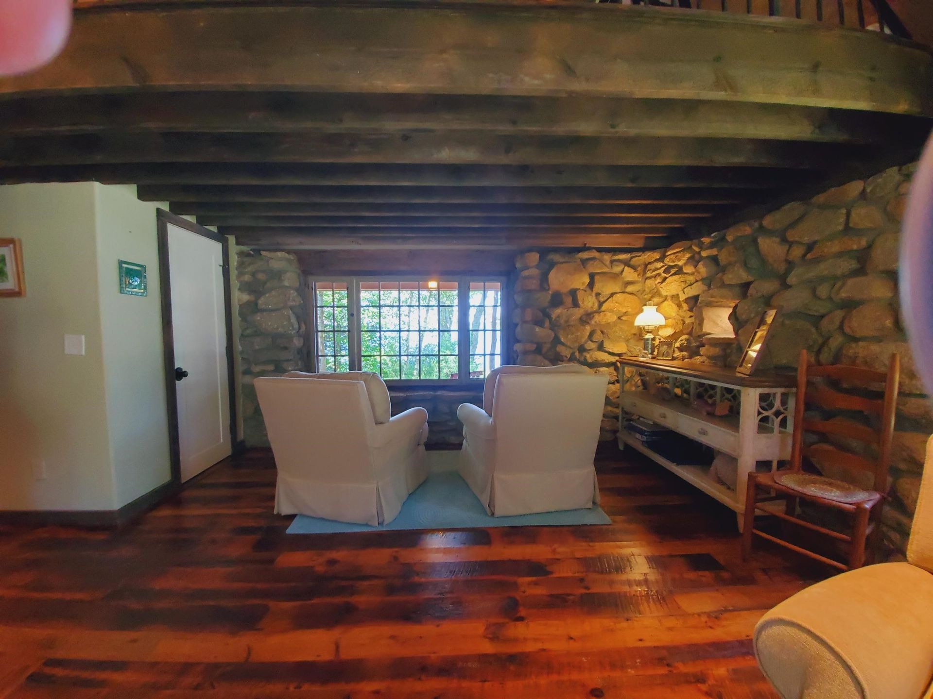 Cozy cabin living room with stone walls, wooden beams, two white armchairs facing a window, and hardwood floors.