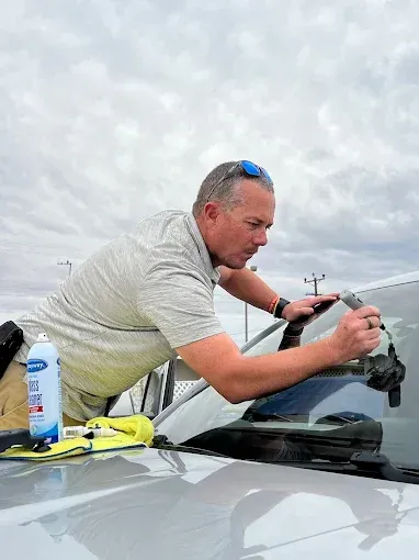 Man applying tint to a car window outdoors, using spray and tools. Cloudy sky.