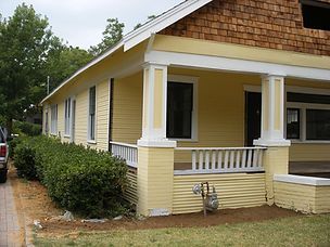 Yellow house with porch, white columns, and brown roof shingles, shrubbery in front.