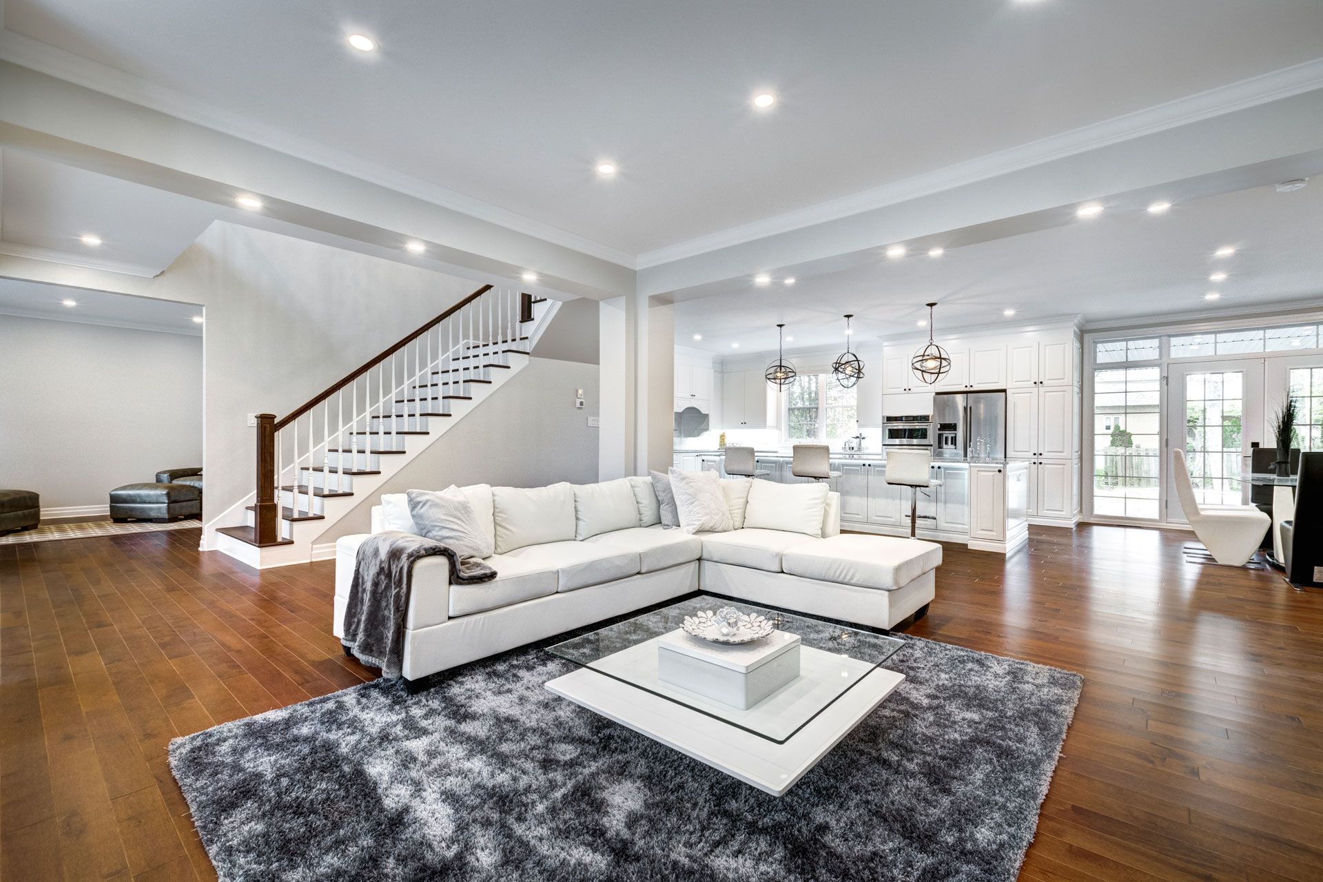 Spacious living room with white sectional sofa, stairs, and open kitchen. Hardwood floors and gray rug.