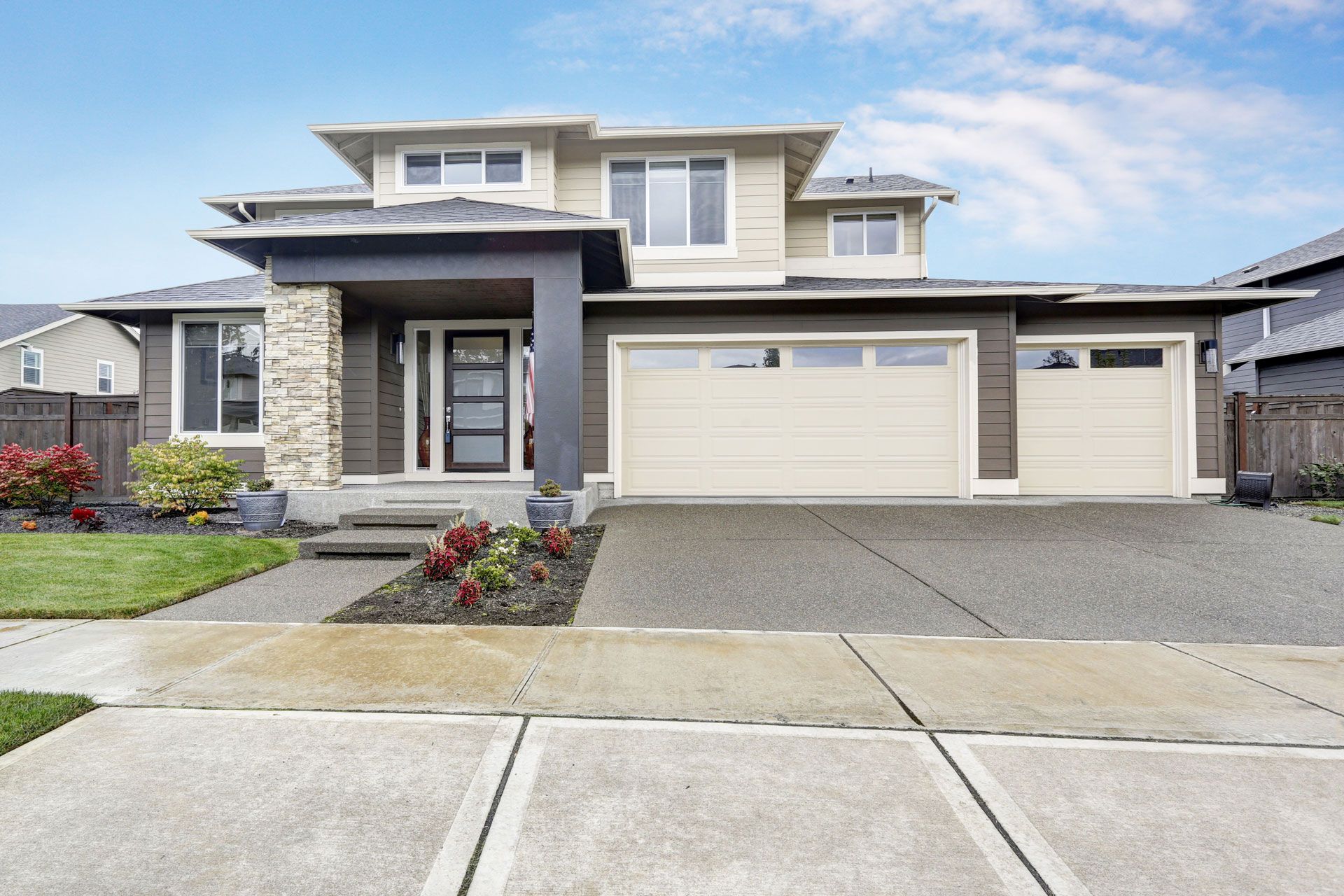 Two-story house with brown exterior, light tan garage doors, stone accent, and concrete driveway on a sunny day.