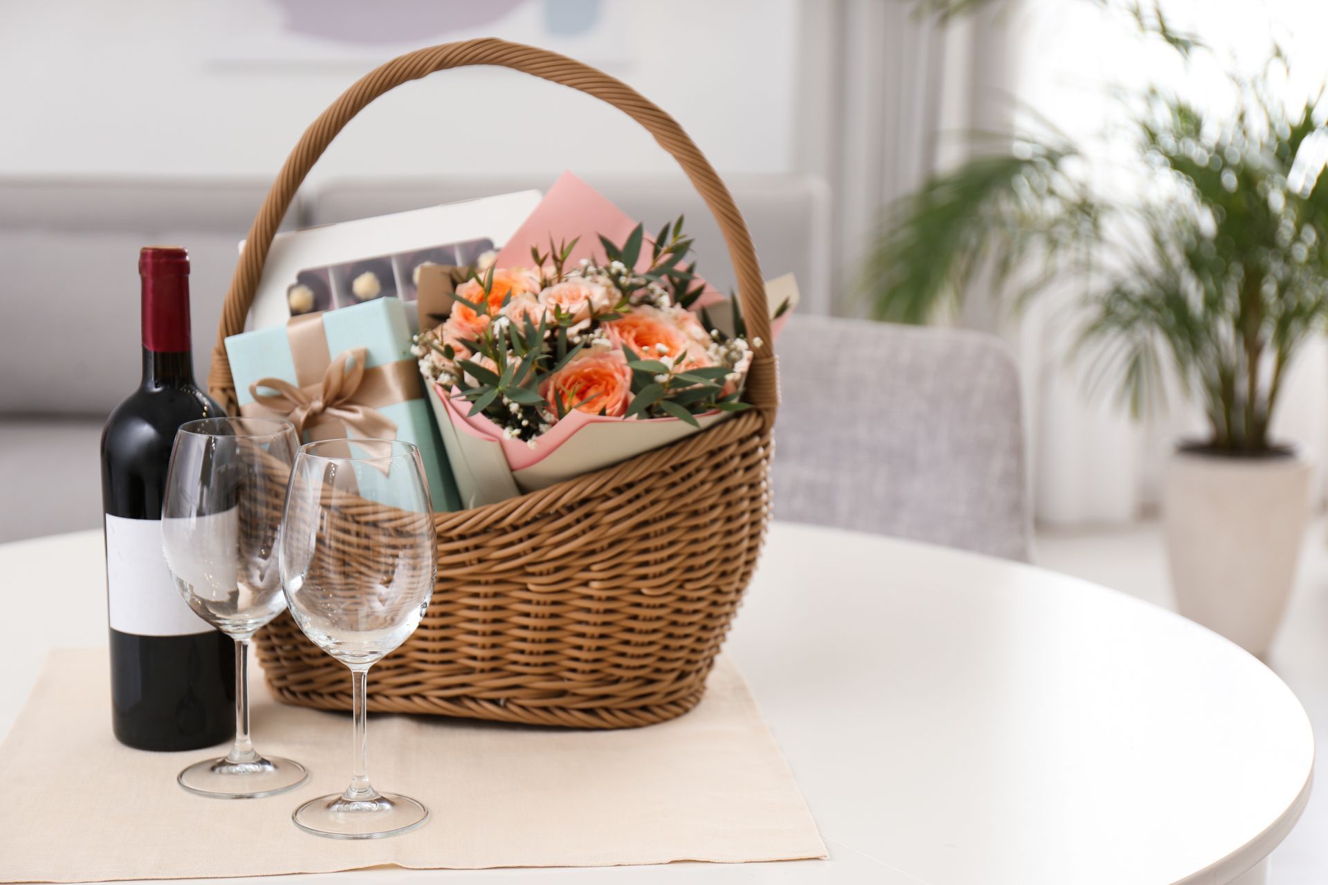 Wicker basket with gifts near bottle of wine and glasses on table indoors.