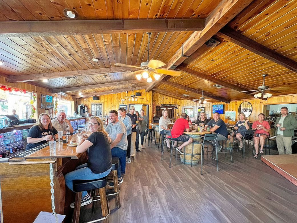 People at a bar with wooden interior. Some sit at the bar while others stand. Ceiling fans and a bar are visible.