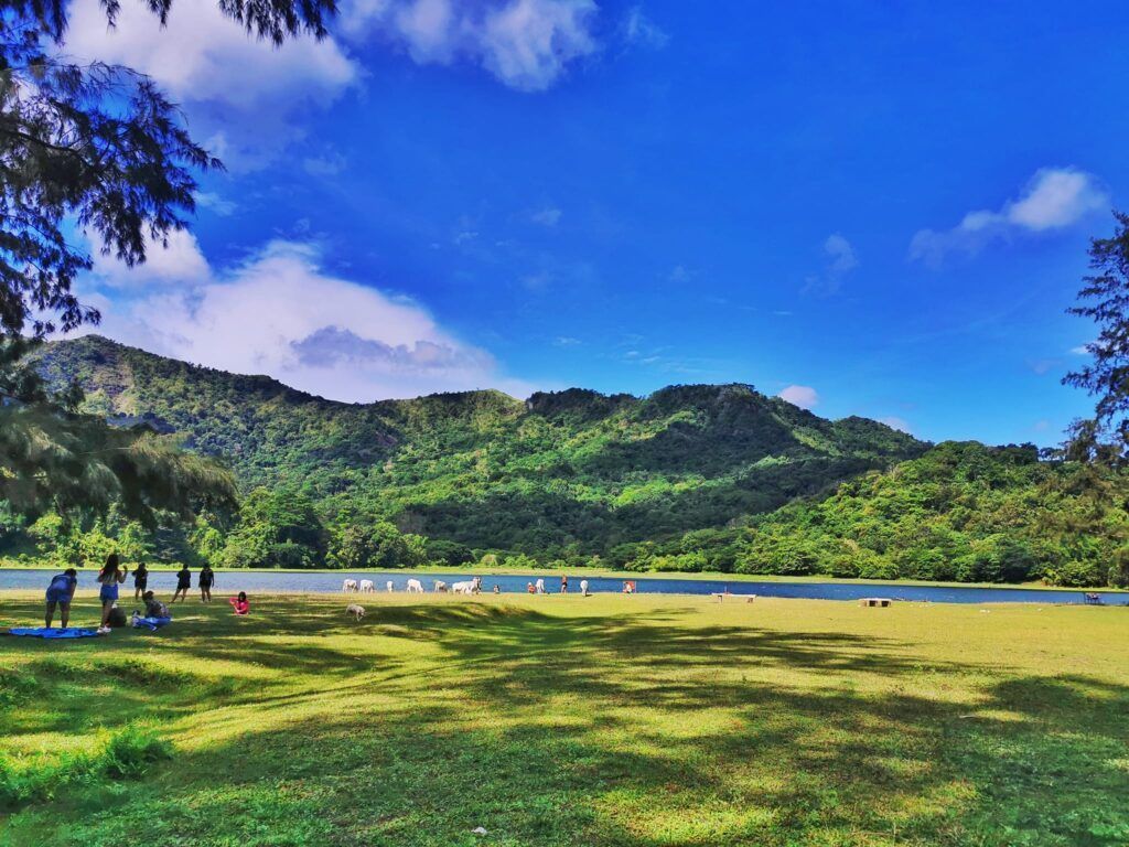 A group of people are sitting on the grass near a lake with mountains in the background.