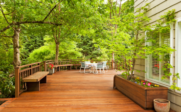 A beautiful photo of a newly built deck surrounded by lush green plants and trees. The deck has flower pots and furniture on it.