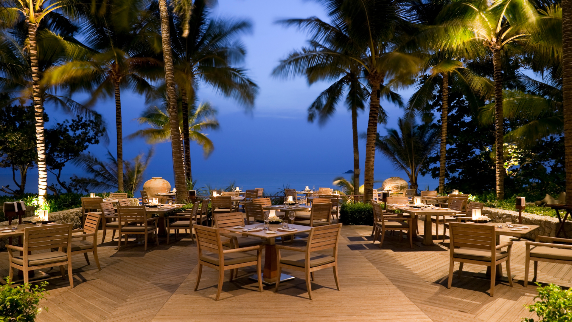 A stunning photo was taken during the evening at a restaurant. In the photo, you can see a commercial deck with several tables and chairs for outside seating. There are trees in the background, and the deck overlooks a lake. This is a beautiful view.