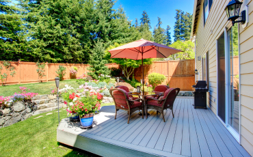 A photo of a patio installed in the backyard of a house. The wooden patio is spacious and has room for a table, umbrella, chairs, flowers, and a grill. The patio overlooks the backyard, which is covered in lush green grass, and a fence guards the perimeter of the backyard. Pine tree branches hang over the fence and act as security to keep the backyard cozy and secure.