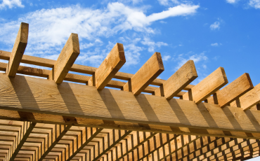 A photo of the roof of a pergola structure. The background is a sunny clear sky with a little bit of white clouds. It is a beautiful day out in this photo. 