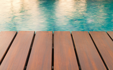 A photo of a newly constructed and installed pool deck. In the photo you see the wooden planks running vertically up the photo and meeting with the water in the pool. The pool water is still and calm. This is a beautiful photo of a pool deck. 