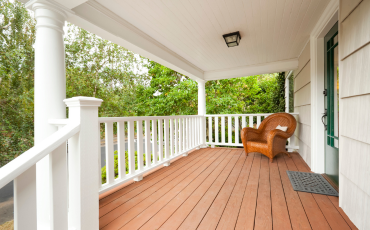 A photo of a porch on the front of a house. The porch has white railings and a white ceiling. The decking boards on the porch are an orange/brown color. There is a comfy chair in the corner of the porch.