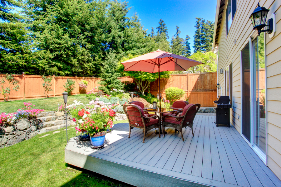 A photo of a patio in the backyard of someone's home. The patio is made out of wood and sits low to the ground. The patio is spacious and has room for furniture, plants and flowers, and even a grill.