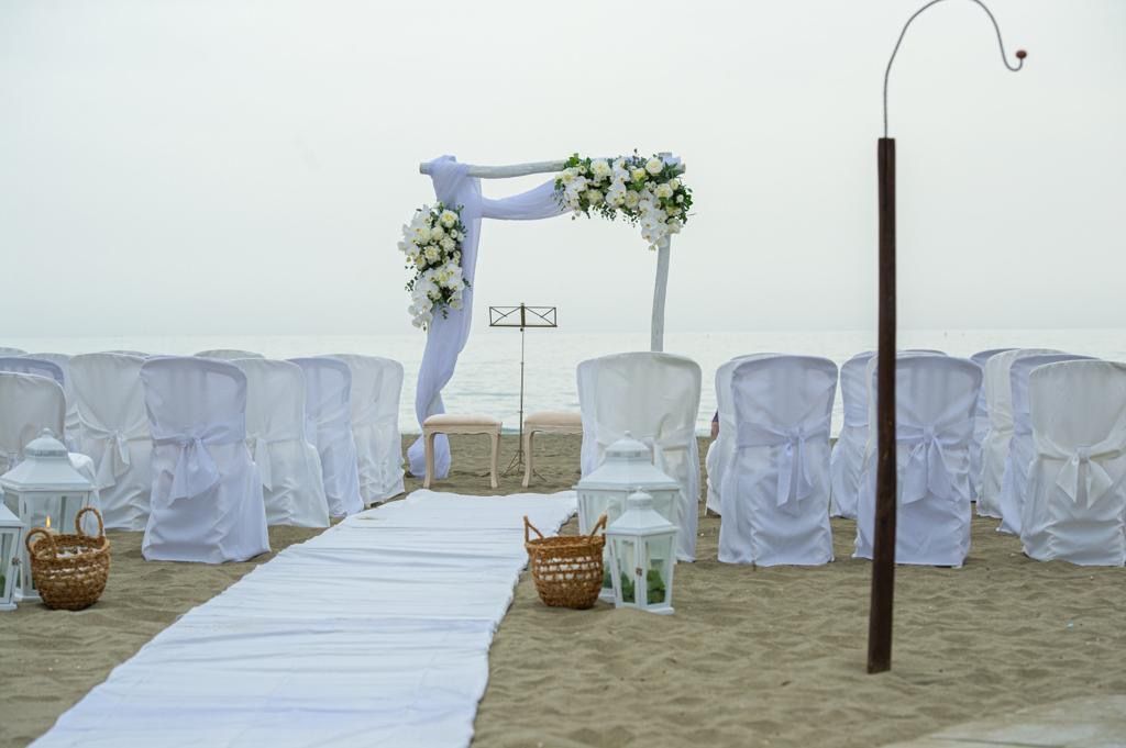 Allestimento per matrimonio sulla spiaggia con sedie bianche, arco e runner lungo la navata, su una spiaggia sabbiosa.