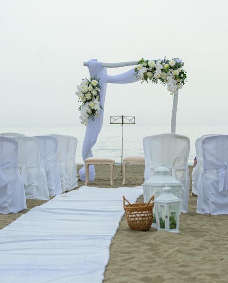 Cerimonia nuziale sulla spiaggia con arco bianco, runner lungo la navata, sedie e composizioni floreali.