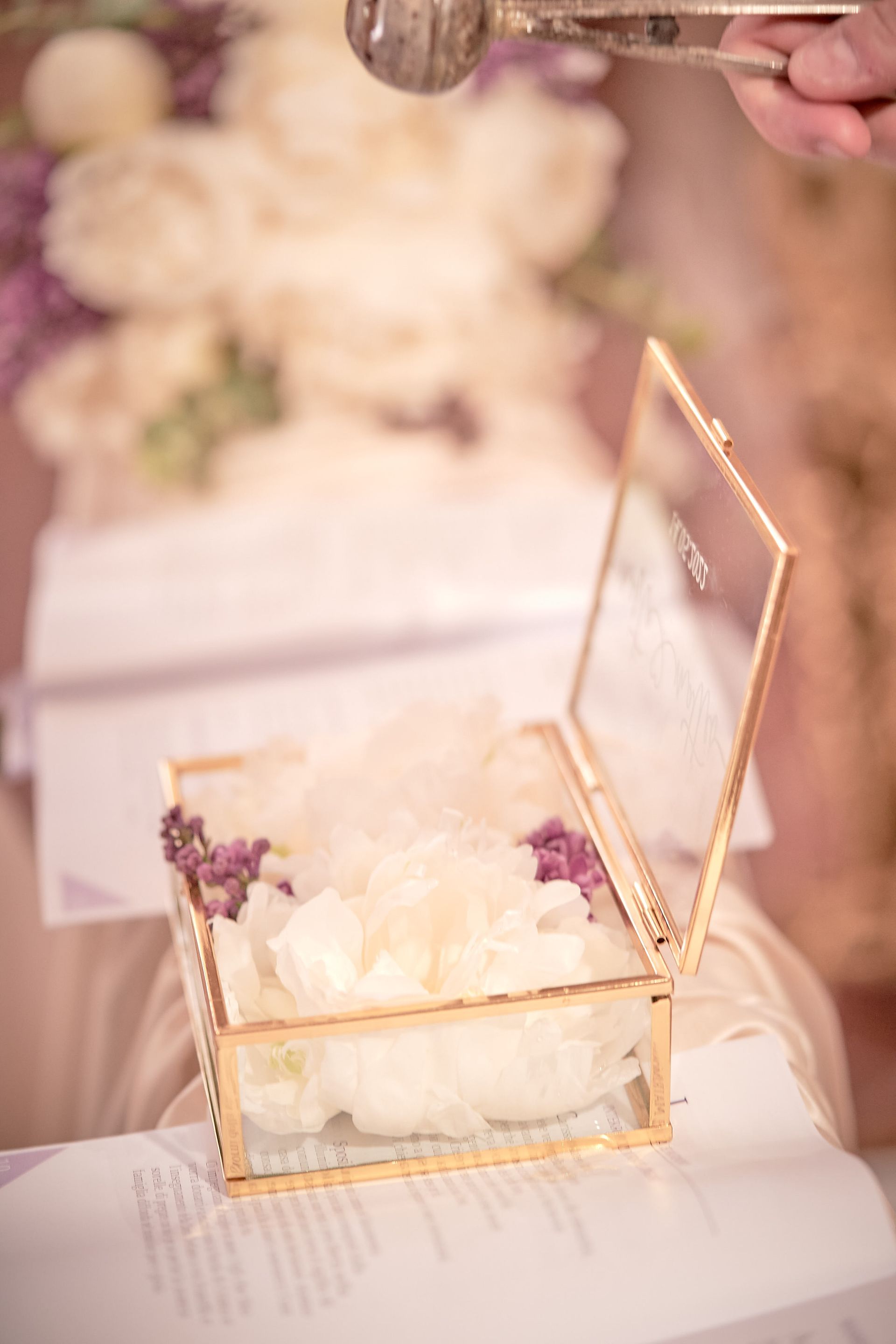 Scatola di vetro aperta con petali di fiori e dettagli viola su un tavolo durante una cerimonia.