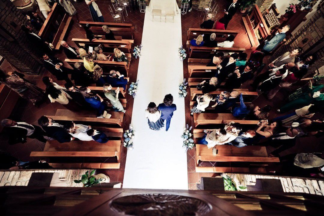 Cerimonia nuziale: la coppia percorre la navata, affiancata dagli invitati seduti su panche di legno.