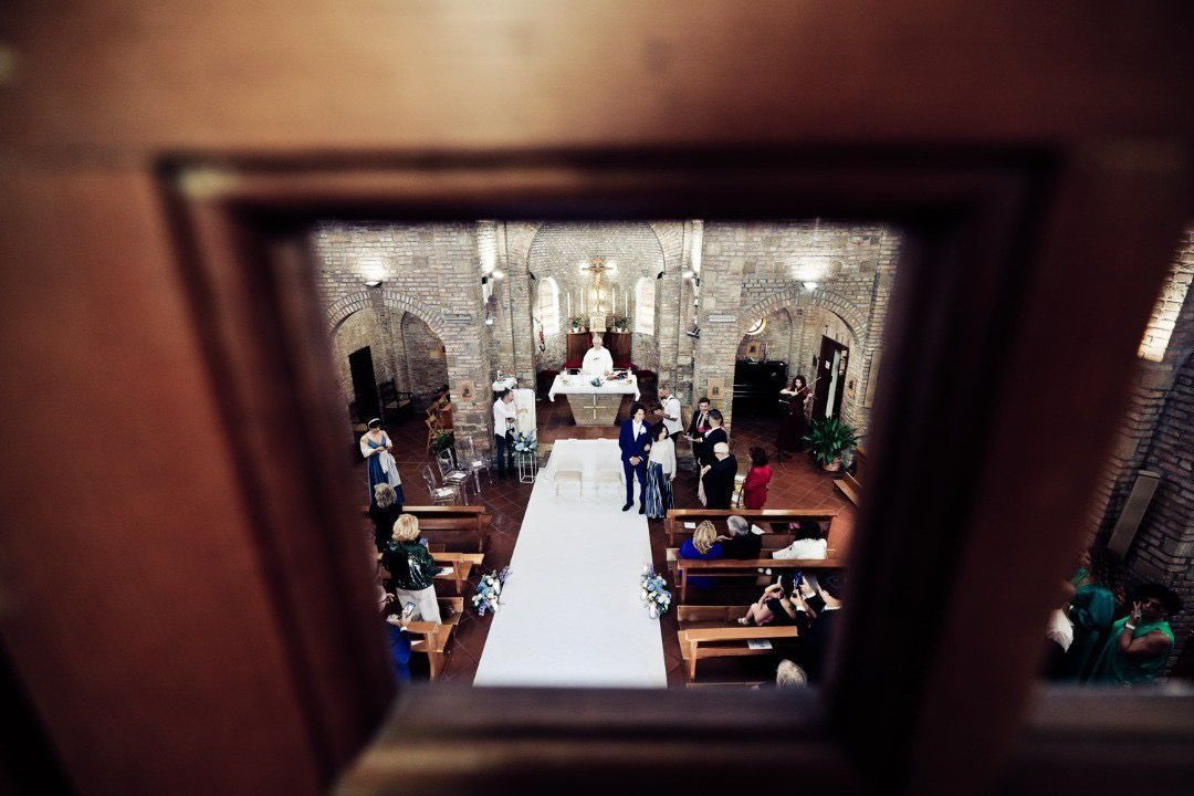 Cerimonia nuziale in chiesa, vista attraverso una porta. Un tappeto bianco conduce all'altare, con gli invitati seduti.