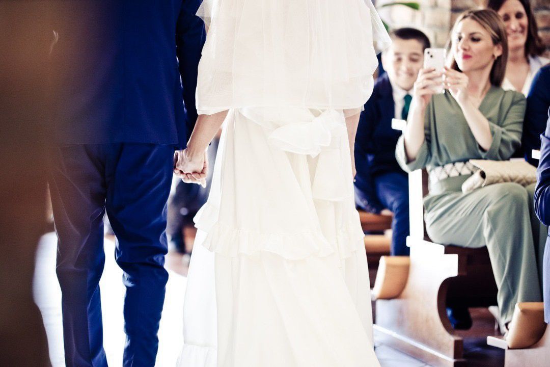 Gli sposi camminano, tenendosi per mano, in una chiesa. Gli ospiti guardano, uno scatta una foto.