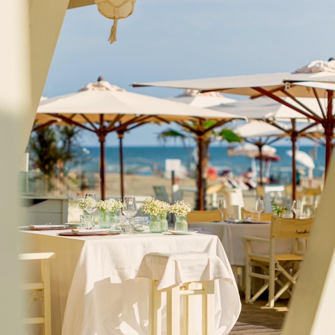 Ristorante sulla spiaggia con tavoli apparecchiati per cenare, oceano e ombrelloni sullo sfondo.