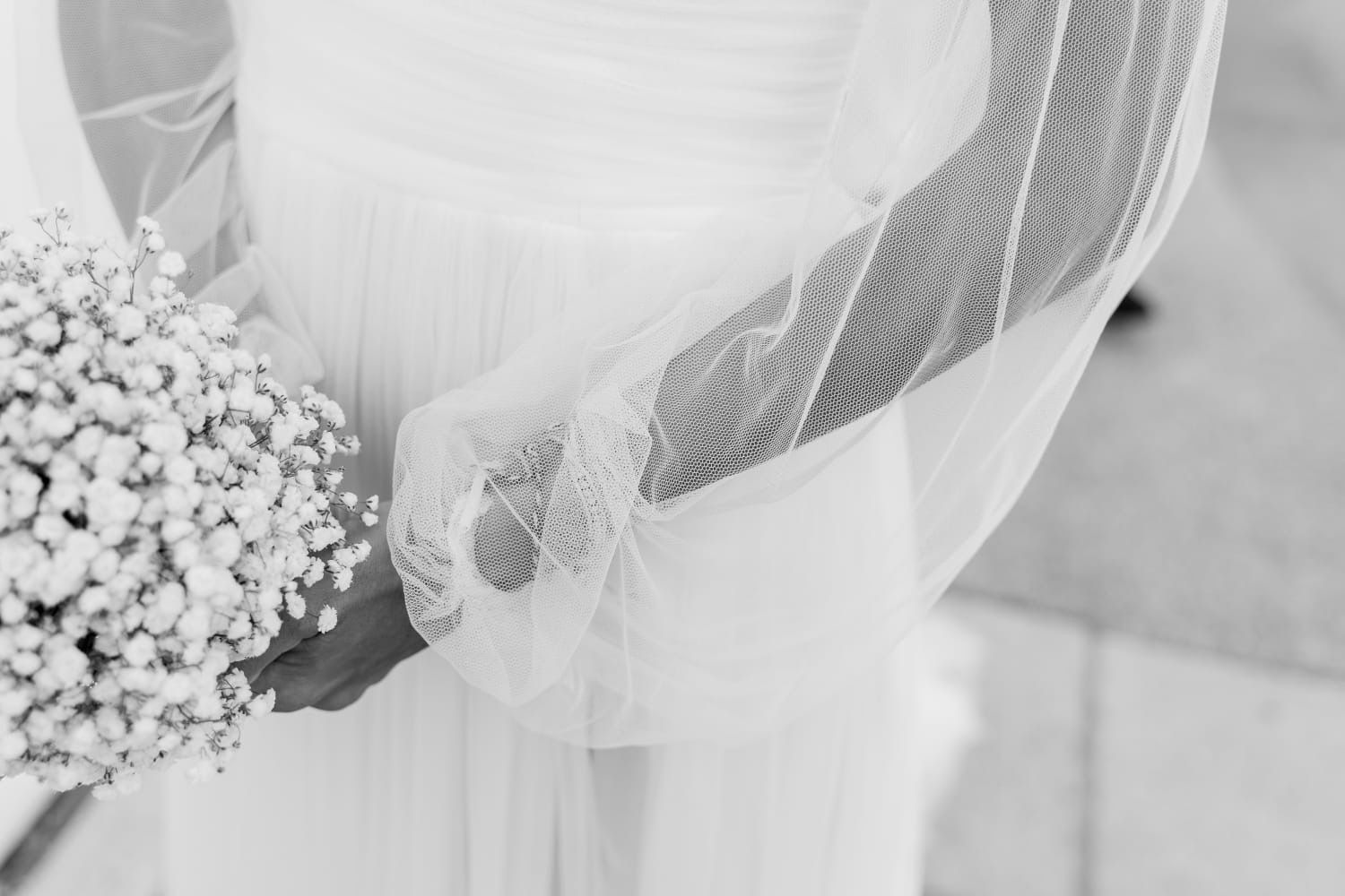 La sposa tiene in mano un bouquet bianco e le braccia sono avvolte in maniche trasparenti bordate di pizzo.