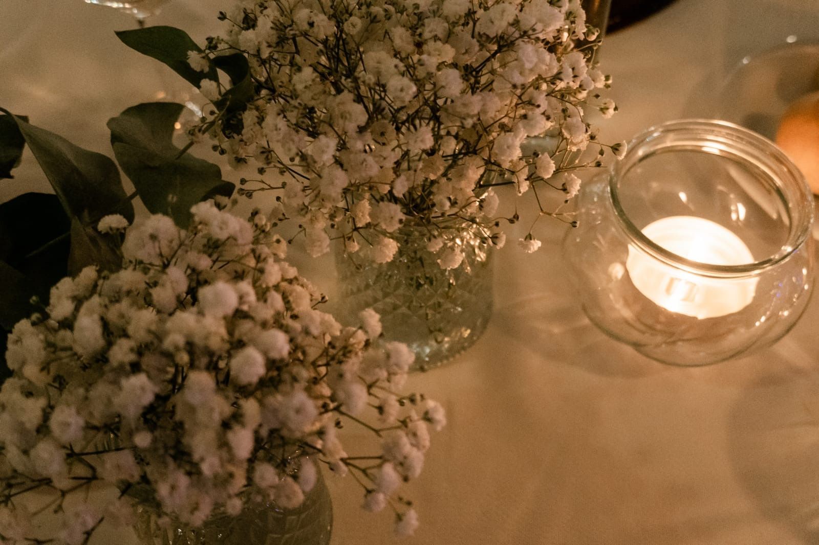 Fiori bianchi di gypsophila in vasi di vetro accanto a una candela accesa in un portacandele di vetro su una tovaglia bianca.