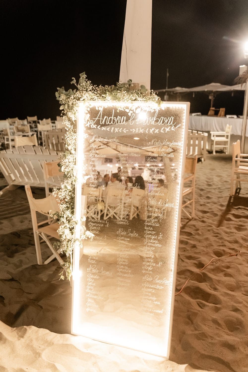 Tabella dei posti a sedere per matrimoni con specchi illuminati su una spiaggia sabbiosa, decorata con verde e luci fatate.