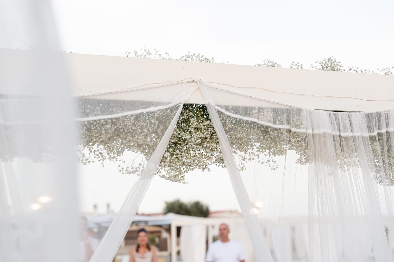 Baldacchino nuziale bianco decorato con gypsophila, tende trasparenti e una coppia sfocata sullo sfondo.
